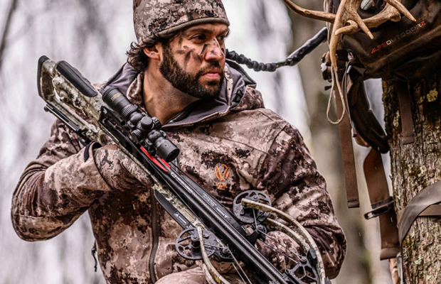 Man holding a crossbow in a treestand.