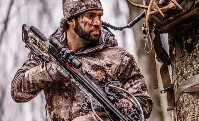 Man holding a crossbow in a treestand.