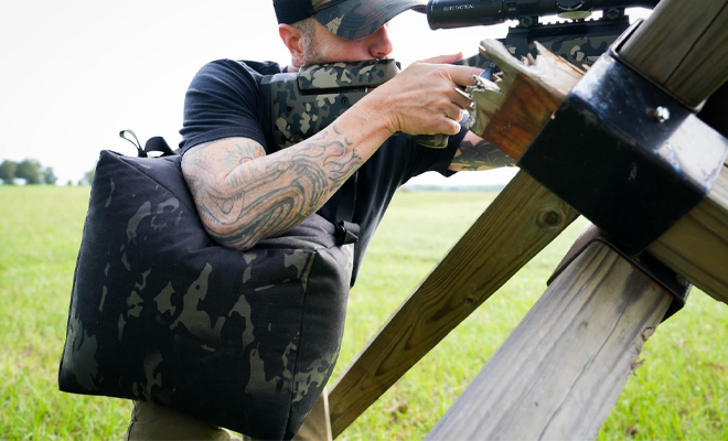 A man shooting a rifle with a sling bag.